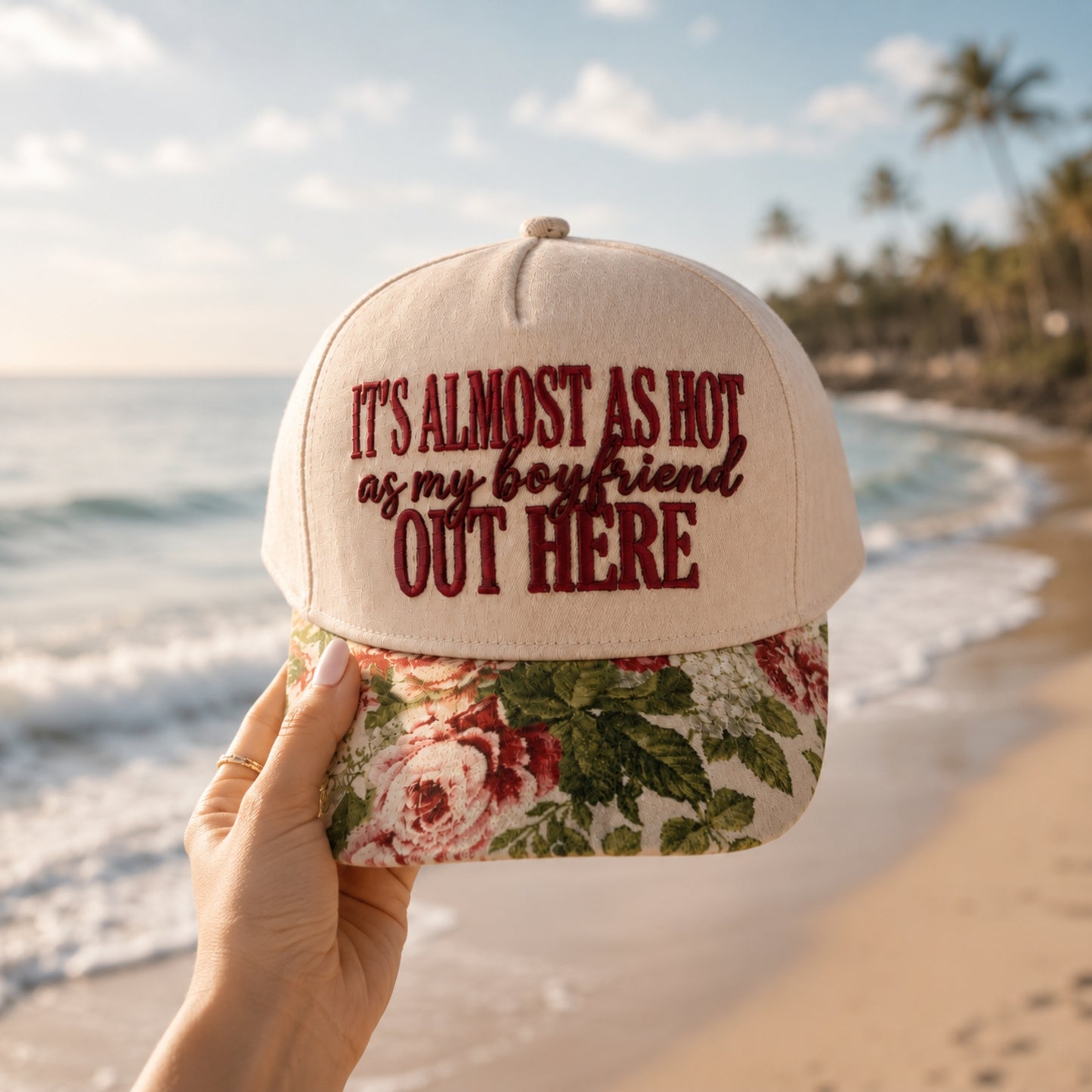 A hand holds a beige baseball cap with dark red embroidered text reading 'IT'S ALMOST AS HOT as my boyfriend OUT HERE' above a floral brim, set against a blurred beach background with ocean waves and palm trees.