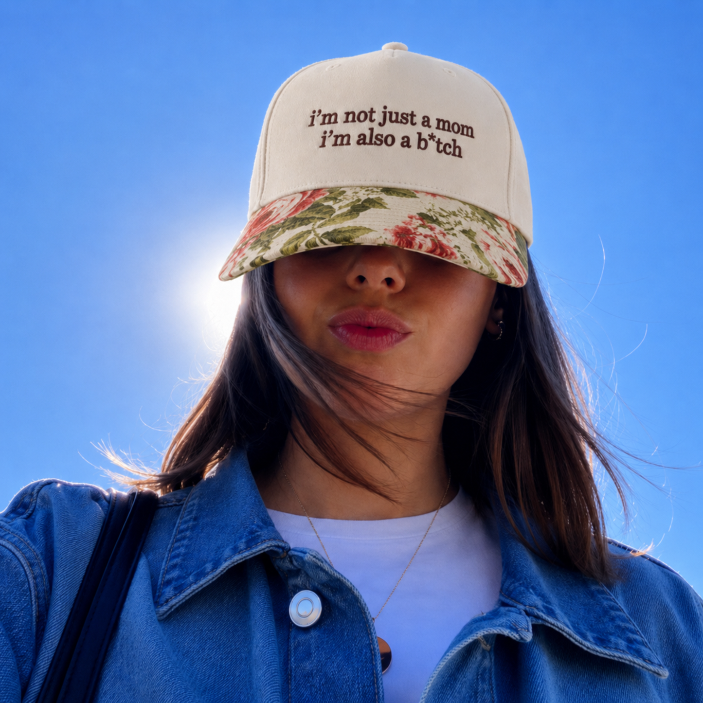 A woman outdoors wearing a cream baseball cap with a floral brim and embroidered text reading 'i'm not just a mom i'm also a b*tch'. The cap is shown against a bright blue sky, with the sun shining behind her.