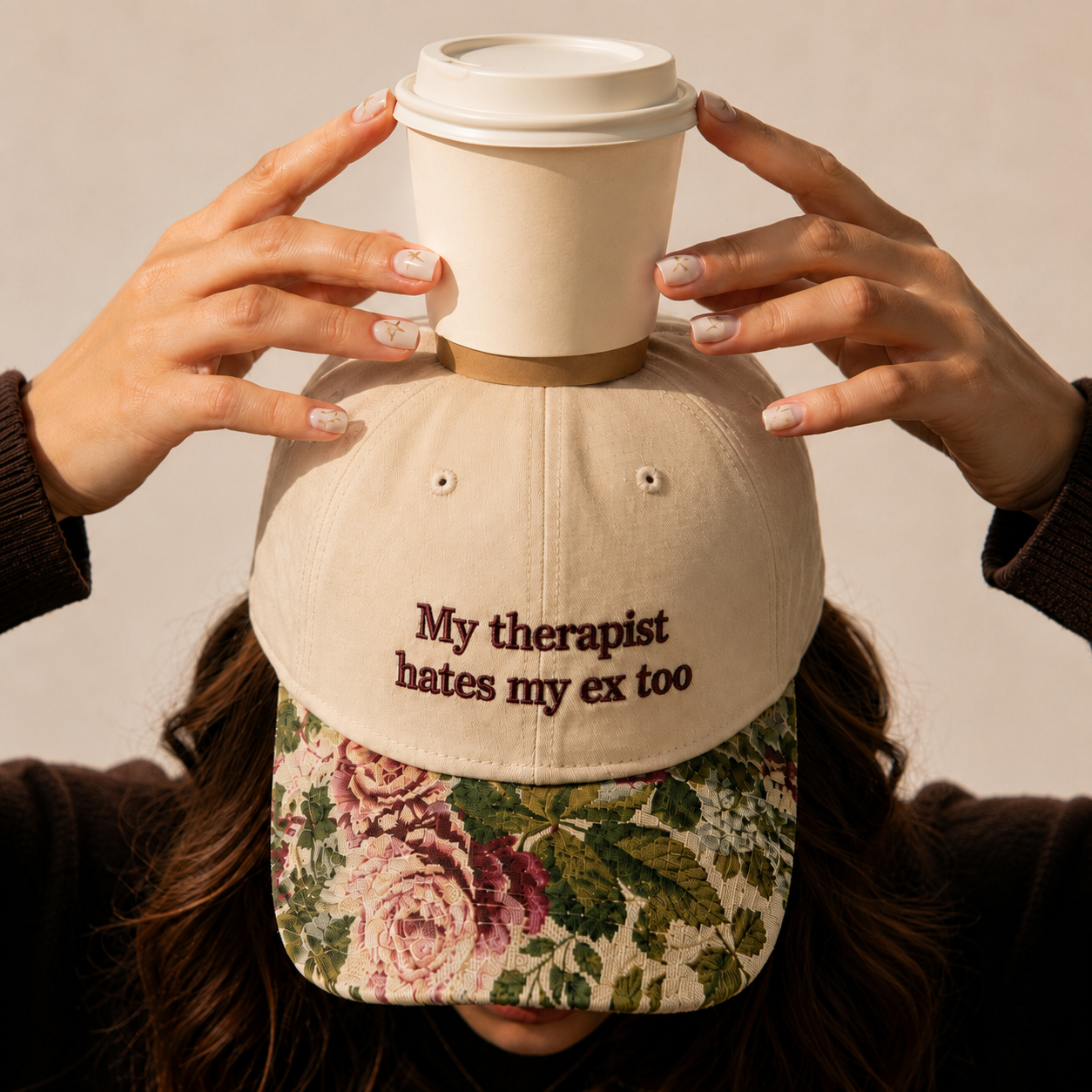 Woman holding a disposable coffee cup on top of a beige baseball cap embroidered with "My therapist hates my ex too" and a vintage floral printed brim.
