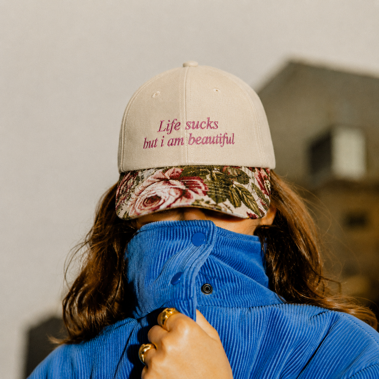 Person wearing a blue jacket and a beige cap embroidered with "Life sucks but i am beautiful" and a floral pattern brim.
