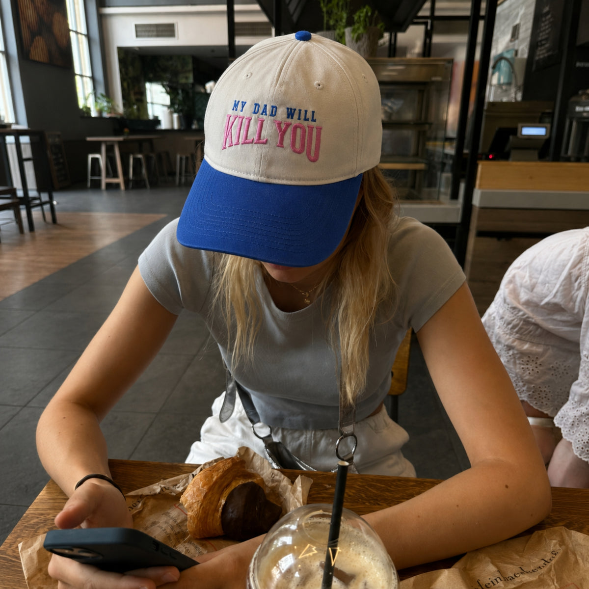 A person sitting at a cafe table wearing a white and blue baseball cap with the text 'MY DAD WILL KILL YOU' in blue and pink embroidery, looking down at a phone.