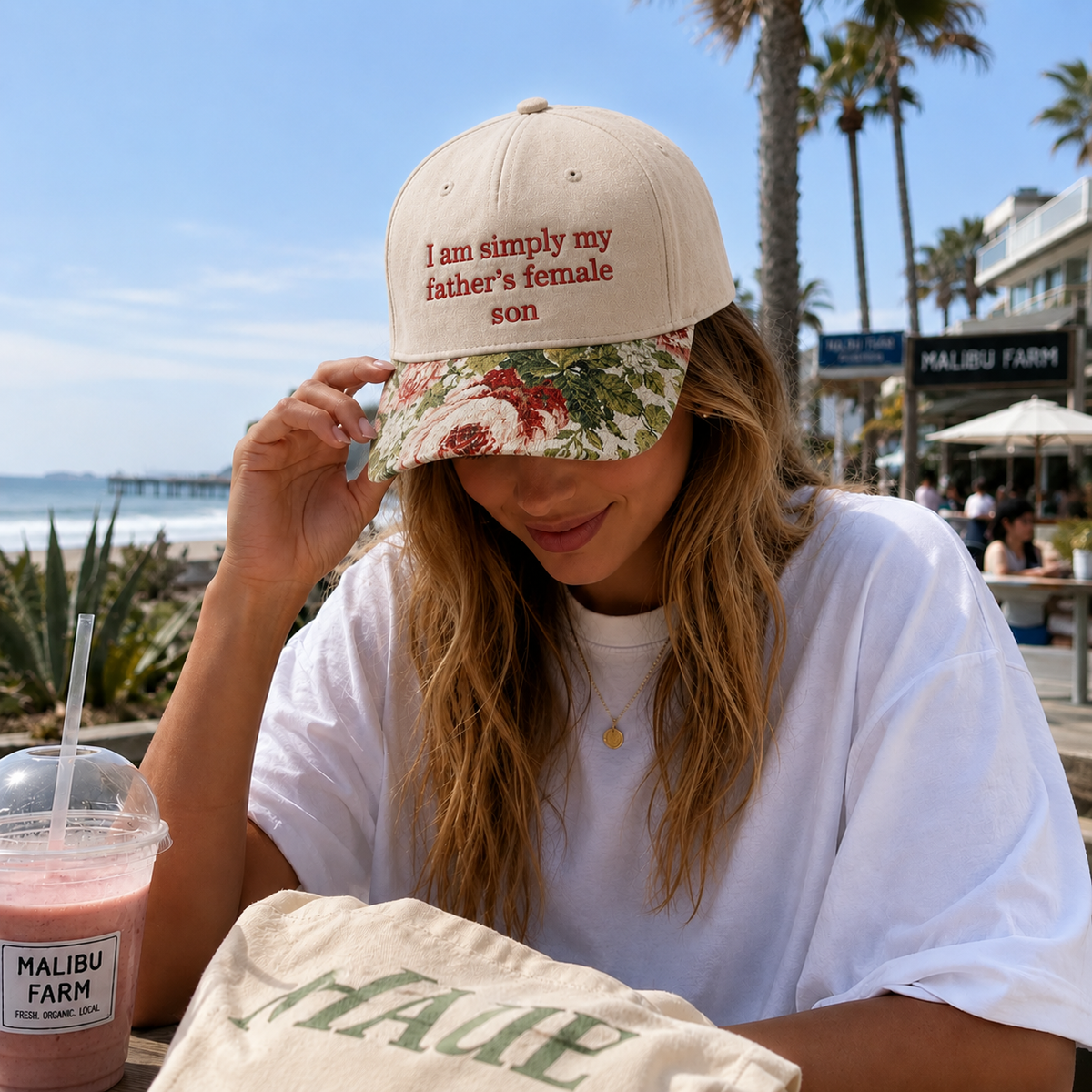 A woman sitting outdoors at a beachside cafe wearing a beige baseball cap with red embroidered text reading 'I am simply my father's female son' and a green floral brim. She is wearing a white t-shirt and a canvas tote bag with 'MALIBU' printed on it, with a pink drink in the foreground.