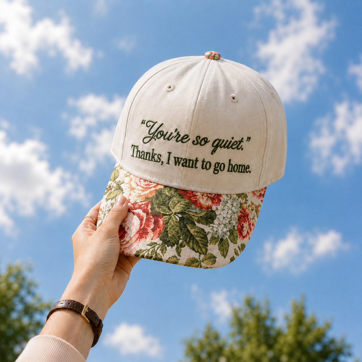 Hand holding up a beige embroidered baseball cap against a blue sky with soft white clouds and green treetops in the background. The cap features dark embroidery reading "You're so quiet." Thanks, I want to go home.` and a vintage floral tapestry pattern on the brim.