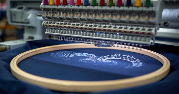 Multi-needle commercial embroidery machine with colorful thread stitching a white design on navy fabric in a hoop