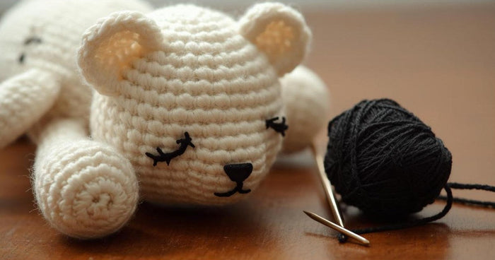 Crocheted amigurumi bear with embroidered sleepy eyes next to black yarn and a tapestry needle