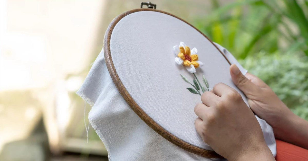 Hands stitching an embroidered flower on fabric held in a wooden embroidery hoop