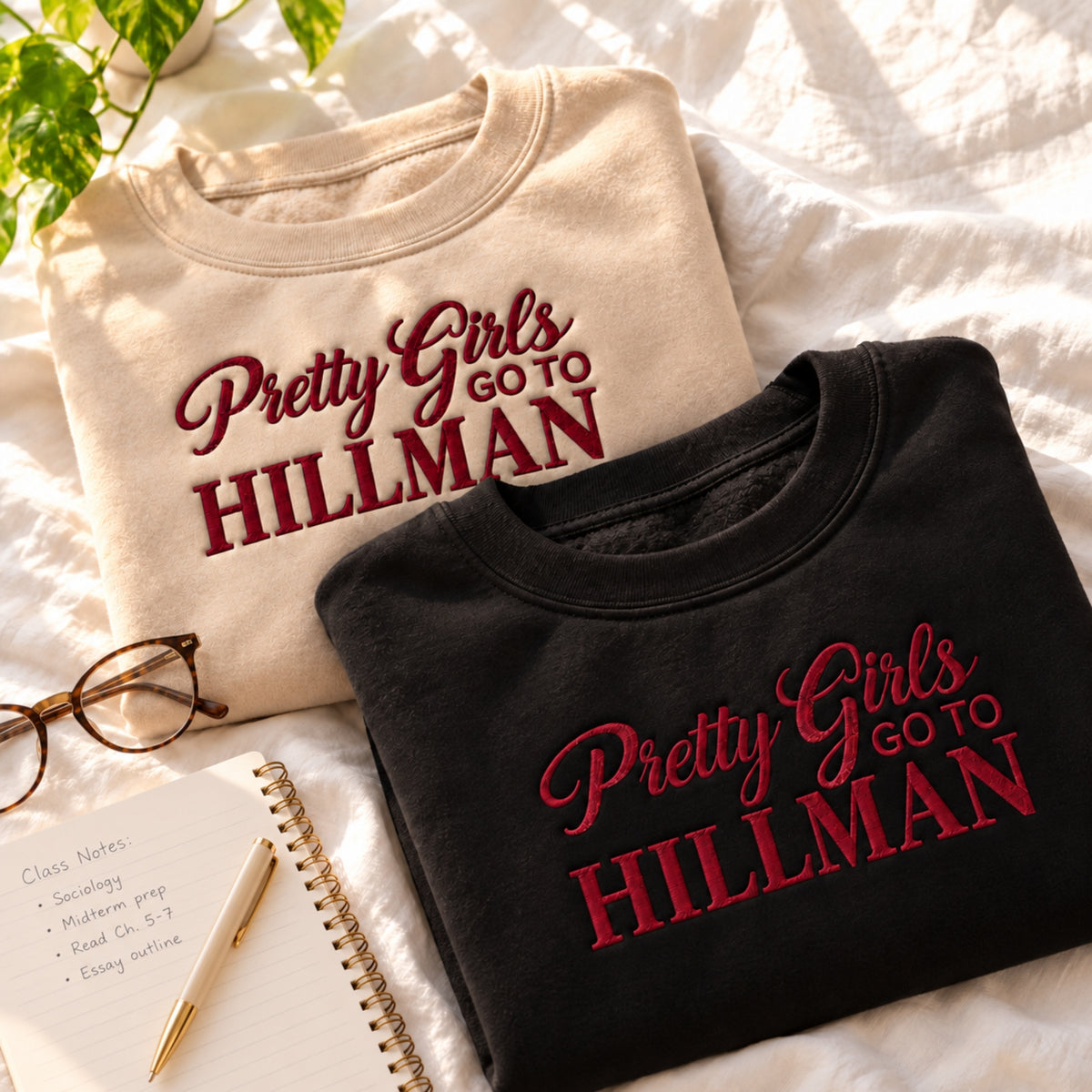 Two folded graphic T-shirts displayed on a white fabric surface with styled props. The top shirt is cream with dark red text reading 'Pretty Girls go to HILLMAN,' and the front shirt is black with matching red text.