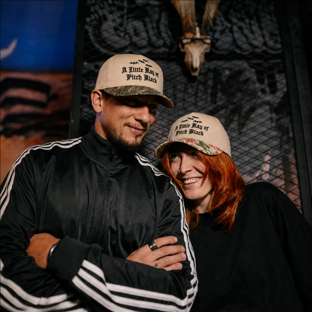 A man and a woman wearing matching beige and camouflage baseball caps with the text 'A Little Way of Pith Black' pose together in front of a rustic, dark-themed wall.