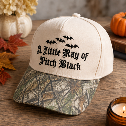 A cream-colored baseball cap with a woodland camouflage brim, featuring black embroidered bats and the text 'A Little Ray of Pitch Black' on the front. The cap is styled on a rustic wooden table with autumn leaves, a candle, and a small pumpkin in the background.