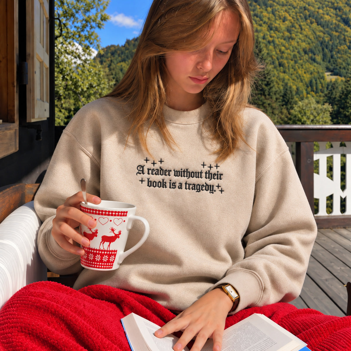 A young woman sits on a wooden deck reading a book while wearing a beige crewneck sweatshirt with a black text graphic and holding a red and white mug. She is wrapped in a red blanket, with a scenic forest background.