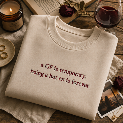 Cream-colored crewneck sweatshirt neatly laid flat on a wooden surface next to a lit candle and a glass of red wine, featuring embroidered text that reads "a GF is temporary, being a hot ex is forever".