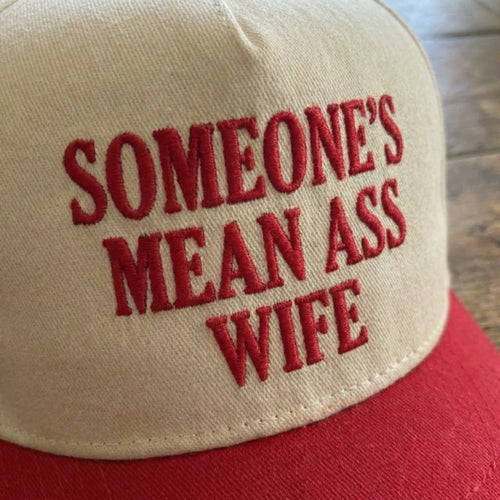 A close-up of a beige baseball cap with red embroidered text reading 'SOMEONE'S MEAN ASS WIFE' and a red brim, photographed on a wooden surface.