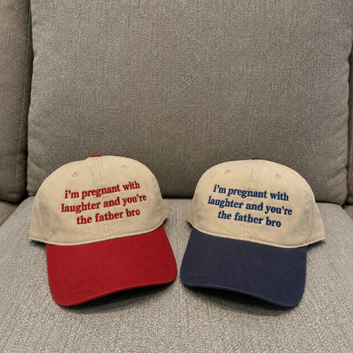 Two baseball caps displayed on a gray upholstered couch. The left cap is beige with a red brim and red embroidered text reading, "I'm pregnant with laughter and you're the father bro.