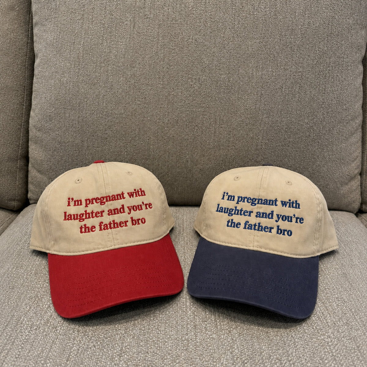 Two baseball caps displayed on a gray upholstered couch. The left cap is beige with a red brim and red embroidered text reading, "I'm pregnant with laughter and you're the father bro.