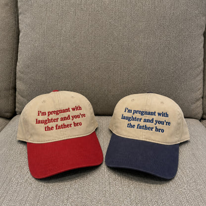 Two baseball caps displayed on a gray upholstered couch. The left cap is beige with a red brim and red embroidered text reading, "I'm pregnant with laughter and you're the father bro.
