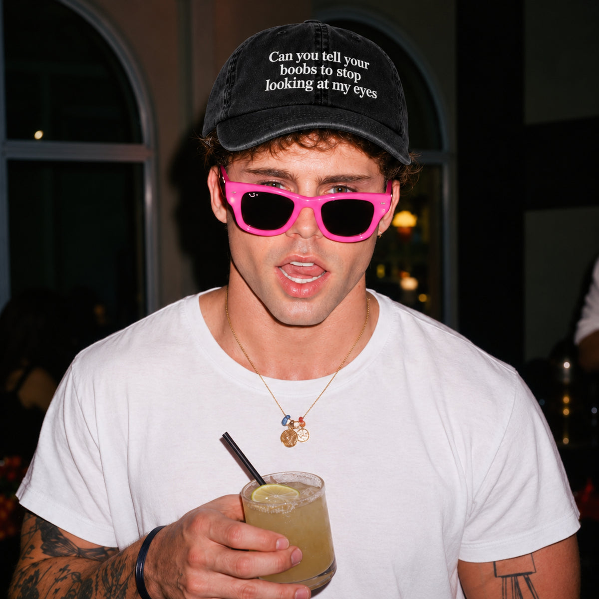 A man wearing a black velvet cap with white embroidered text, hot pink sunglasses, and a white t-shirt, holding a drink in a dimly lit indoor setting.