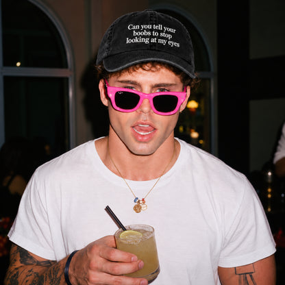 A man wearing a black velvet cap with white embroidered text, hot pink sunglasses, and a white t-shirt, holding a drink in a dimly lit indoor setting.