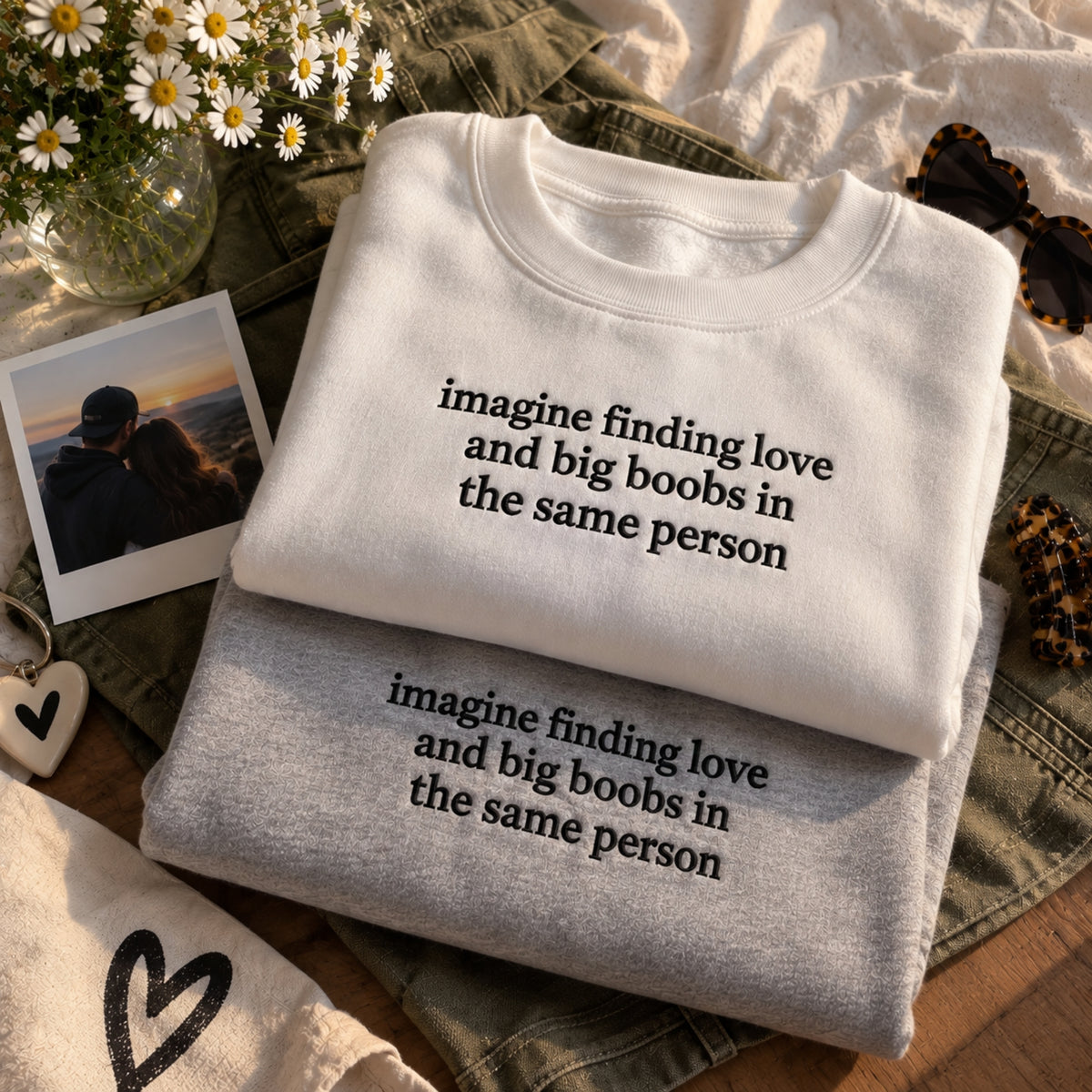 Two folded crewneck sweatshirts stacked on top of each other, one white and one heather gray, both printed with the phrase 'imagine finding love and big boobs in the same person' in black lowercase text. The sweatshirts are styled in a flat lay with denim shorts, daisies in a glass vase, a Polaroid photo, and sunglasses.
