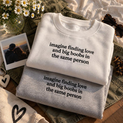 Two folded crewneck sweatshirts stacked on top of each other, one white and one heather gray, both printed with the phrase 'imagine finding love and big boobs in the same person' in black lowercase text. The sweatshirts are styled in a flat lay with denim shorts, daisies in a glass vase, a Polaroid photo, and sunglasses.