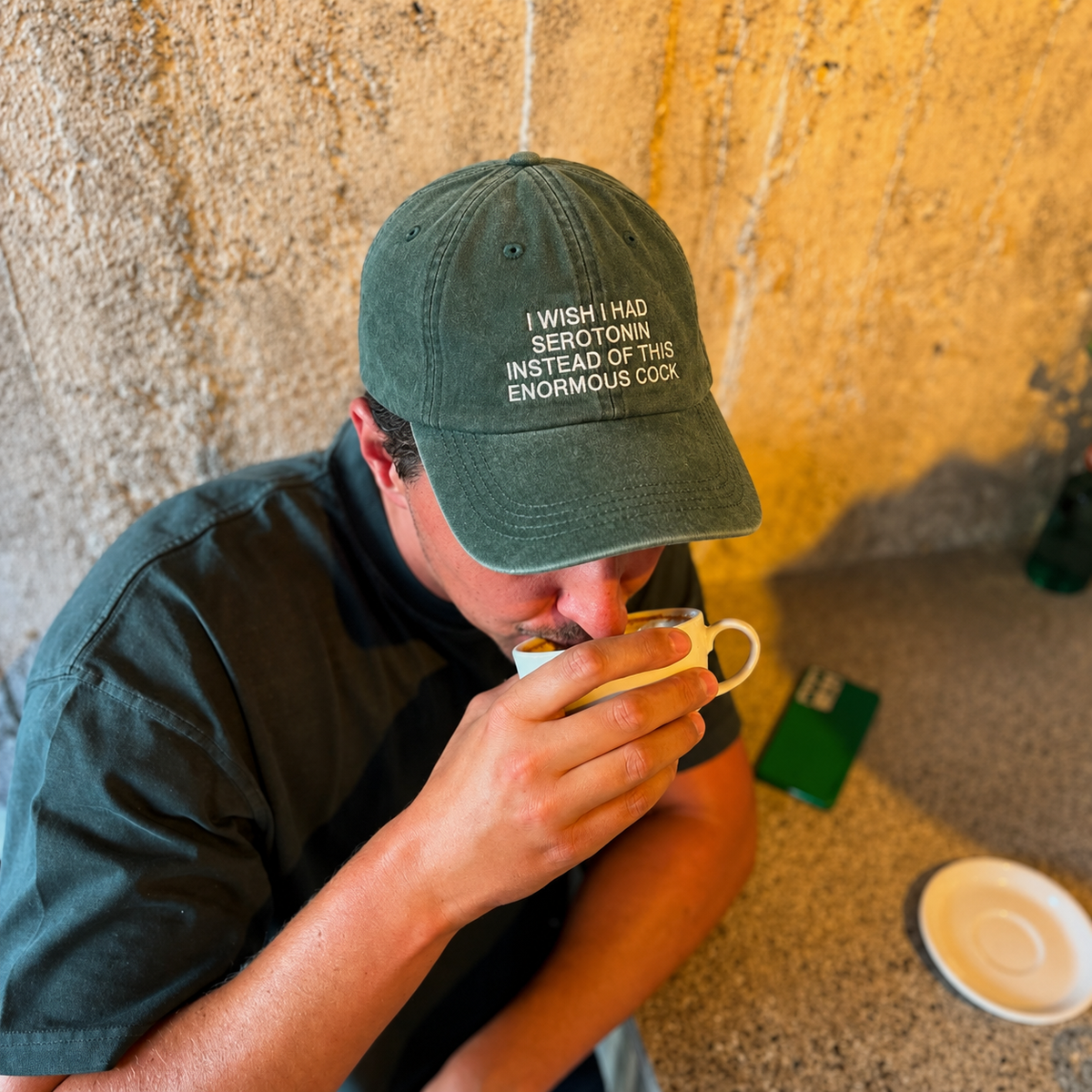 A man wearing a dark green embroidered baseball cap with the text 'I WISH I HAD SEROTONIN INSTEAD OF THIS ENORMOUS GOOP' and drinking from a small white cup in a warm, rustic setting.