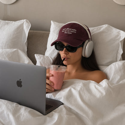 A woman lying in bed under white sheets wears a burgundy baseball cap with white text, white over-ear headphones, and black sunglasses while drinking a pink smoothie and using a gray laptop.