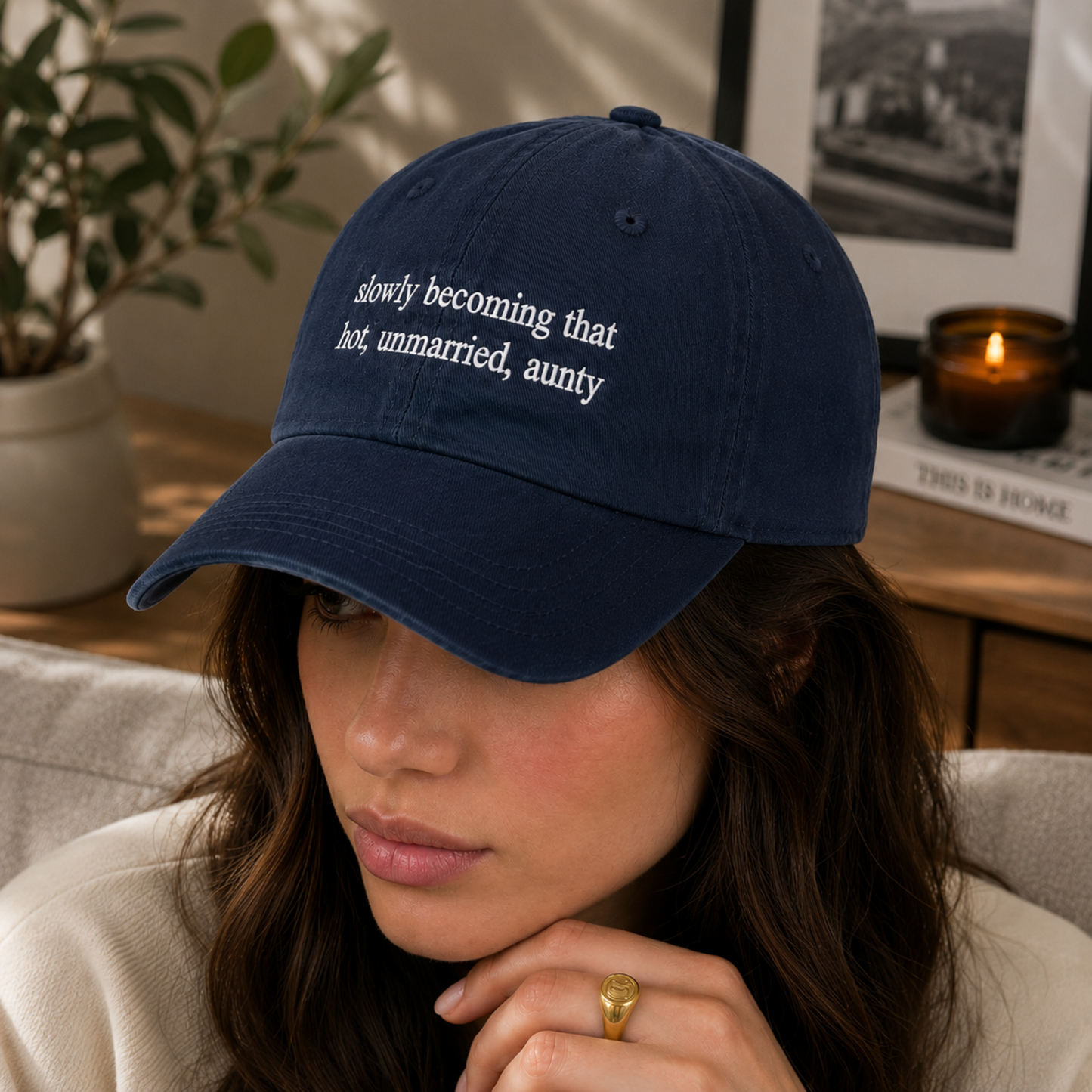 A woman wearing a navy blue baseball cap with white embroidered text that reads 'slowly becoming that hot, unmarried, aunty'. The photo is taken in a cozy indoor setting with a lit candle, framed picture, and potted plant in the background.