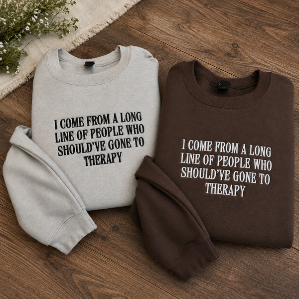 Two folded crewneck sweatshirts displayed on a wooden surface: one light heather gray with black text reading 'I COME FROM A LONG LINE OF PEOPLE WHO SHOULD'VE GONE TO THERAPY' and one dark brown with white text reading the same phrase. A small floral prop and woven fabric corner are visible at the top left.