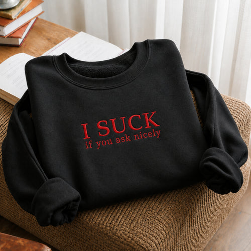 A black crewneck sweatshirt with red embroidered text reading 'I SUCK if you ask nicely' is folded and styled on a brown woven chair next to an open book on a wooden table.