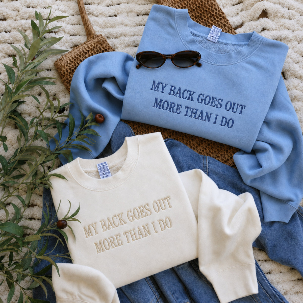 Two folded crewneck sweatshirts with the text 'MY BACK GOES OUT MORE THAN I DO' are arranged on a cream knit blanket with styling props. The top sweatshirt is light blue with dark blue lettering, styled with sunglasses and a woven bag.