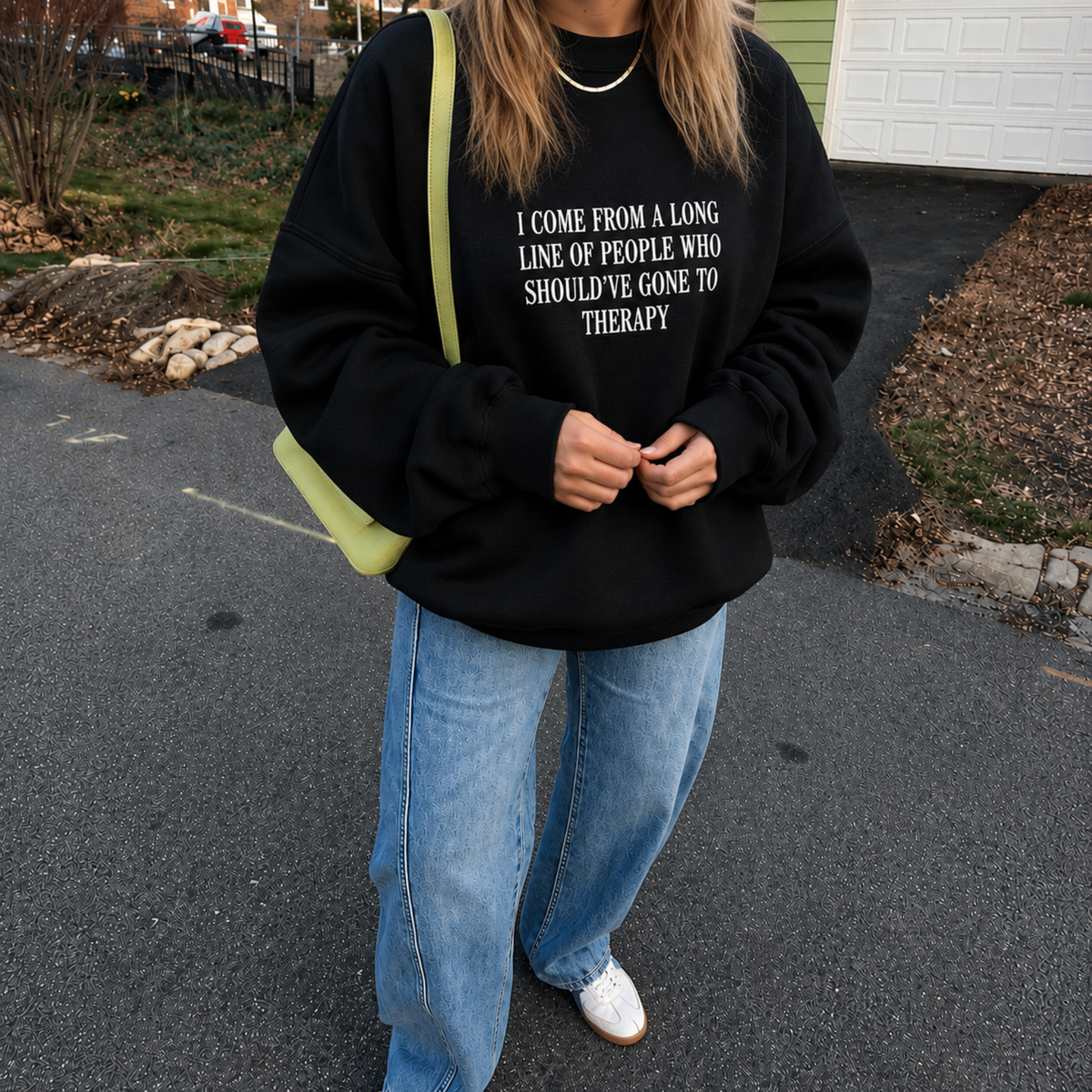 A person wearing a black oversized sweatshirt with white text that reads 'I COME FROM A LONG LINE OF PEOPLE WHO SHOULDN'T HAVE GONE TO THERAPY', paired with light blue jeans and a lime green shoulder bag, standing on an asphalt driveway.