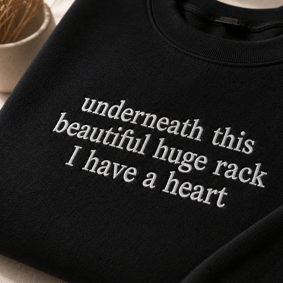 A close-up of a black crewneck sweatshirt with white embroidered text reading 'underneath this beautiful huge rack I have a heart'.