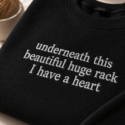 A close-up of a black crewneck sweatshirt with white embroidered text reading 'underneath this beautiful huge rack I have a heart'.