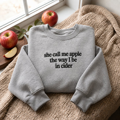 A light gray crewneck sweatshirt with the text 'she call me apple the way I be in cider' printed in black on the front. The sweatshirt is styled in a cozy setting on a beige knit blanket, with red apples and a window in the background.