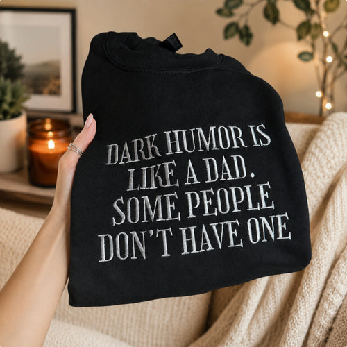 A hand holds a black crewneck sweatshirt with white text that reads 'DARK HUMOR IS LIKE A DAD. SOME PEOPLE DON'T HAVE ONE'.