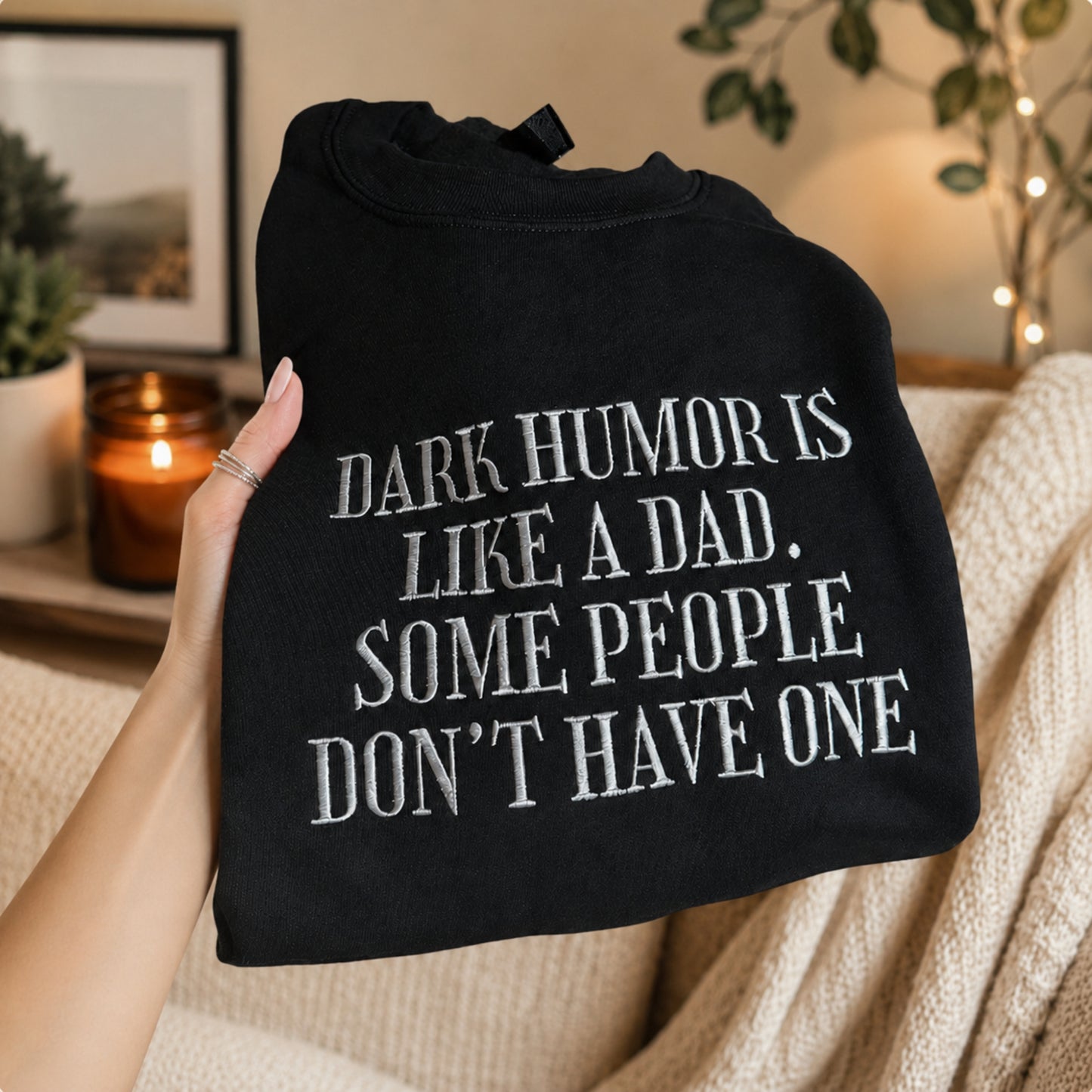 A hand holds a black crewneck sweatshirt with white text that reads 'DARK HUMOR IS LIKE A DAD. SOME PEOPLE DON'T HAVE ONE'.