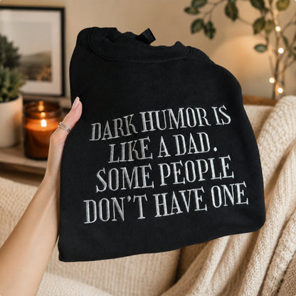 A hand holds a black crewneck sweatshirt with white text that reads 'DARK HUMOR IS LIKE A DAD. SOME PEOPLE DON'T HAVE ONE'.