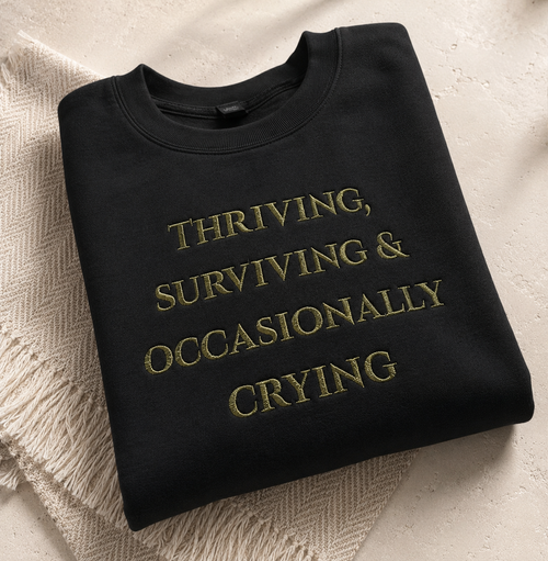 A folded black crewneck sweatshirt with gold embroidered text reading 'THRIVING, SURVIVING & OCCASIONALLY CRYING' on a textured beige surface with a cream knit blanket underneath.