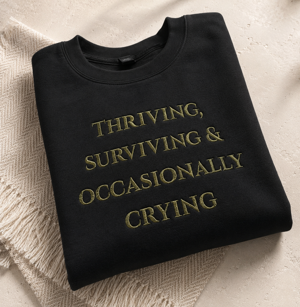 A folded black crewneck sweatshirt with gold embroidered text reading 'THRIVING, SURVIVING & OCCASIONALLY CRYING' on a textured beige surface with a cream knit blanket underneath.