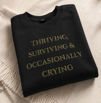 A folded black crewneck sweatshirt with gold embroidered text reading 'THRIVING, SURVIVING & OCCASIONALLY CRYING' on a textured beige surface with a cream knit blanket underneath.