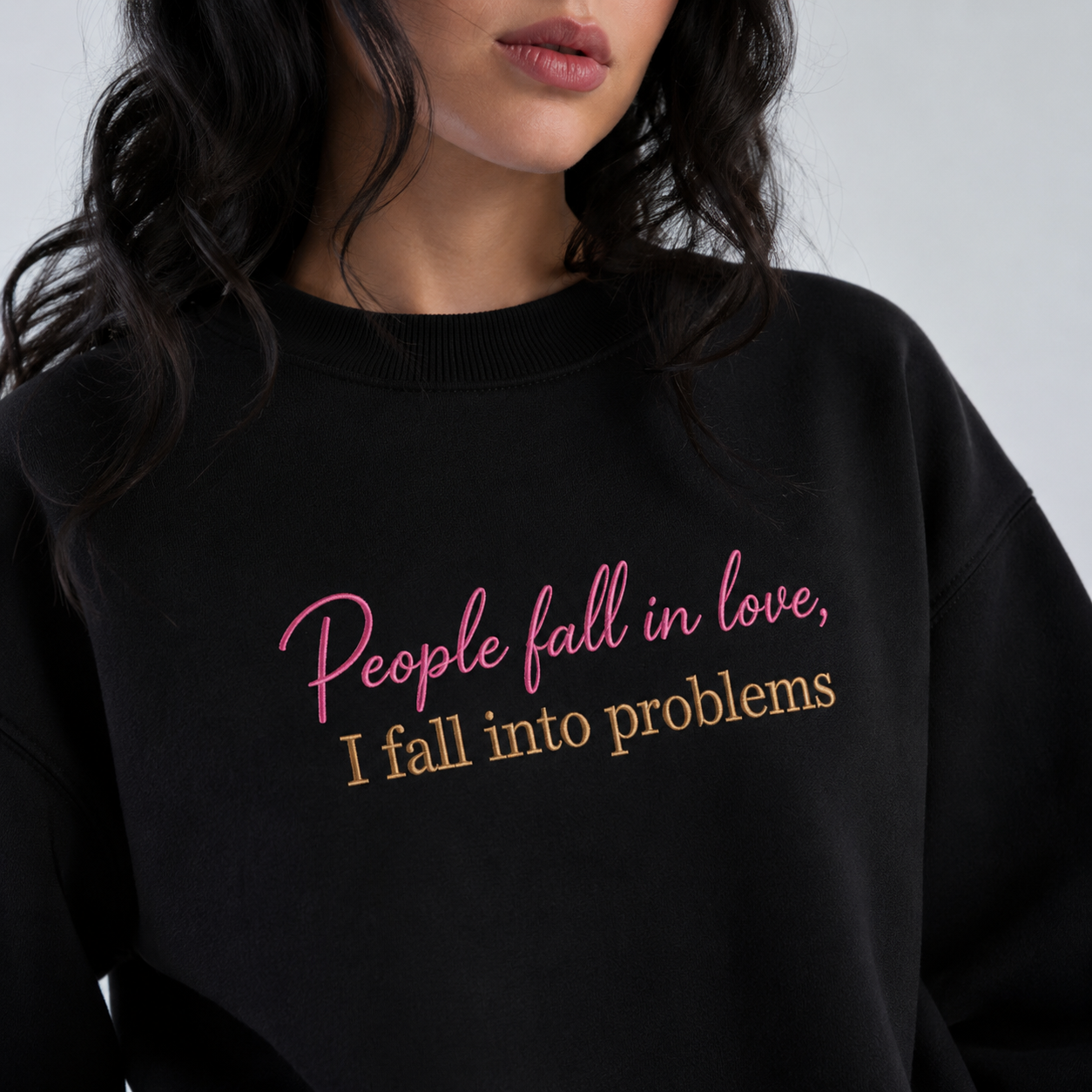 Woman wearing a black crewneck sweatshirt embroidered with pink cursive text reading "People fall in love," above beige block lettering reading "I fall into problems."