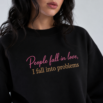 Woman wearing a black crewneck sweatshirt embroidered with pink cursive text reading "People fall in love," above beige block lettering reading "I fall into problems."