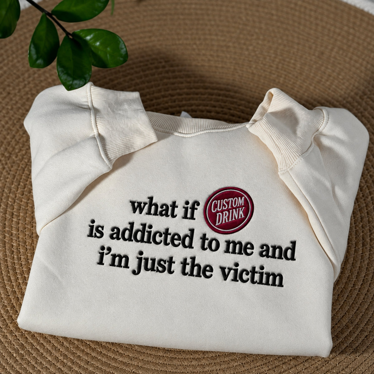 An off-white crewneck sweatshirt with black text reading 'what if is addicted to me and i'm just the victim' and a red circular badge that says 'CUSTOM DRINK', displayed on a brown woven mat with green leaves in the corner.