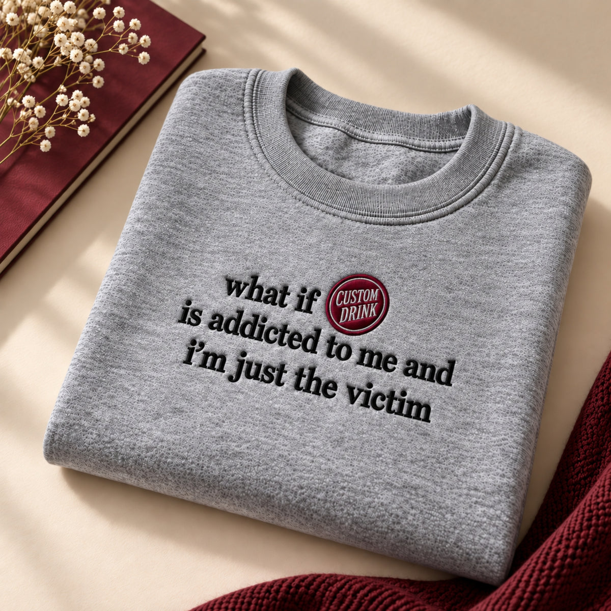 A folded heather gray crewneck sweatshirt with black text reading 'what if is addicted to me and I'm just the victim' and a circular red-and-black 'CUSTOM DRINK' logo, styled on a beige surface with a book, dried flowers, and a red knit fabric.