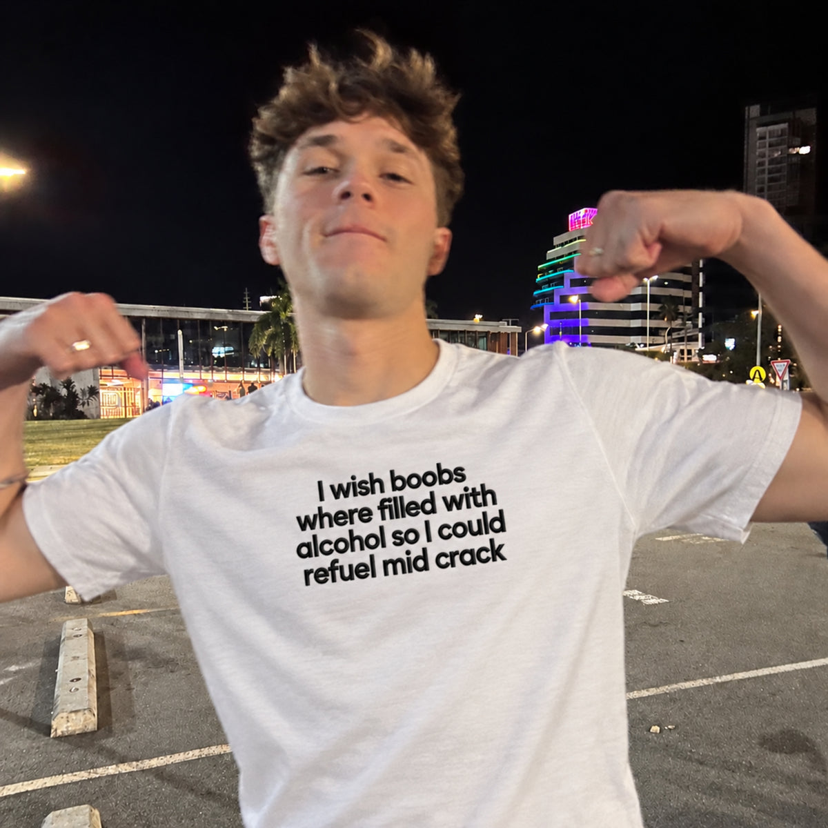 A young man posing in a parking lot at night wearing a white graphic t-shirt with black text that reads 'I wish boobs where filled with alcohol so I could refuel mid crack'.