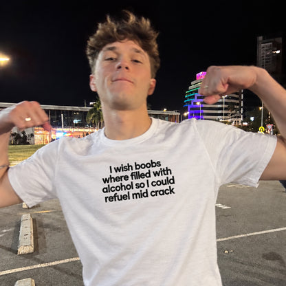 A young man posing in a parking lot at night wearing a white graphic t-shirt with black text that reads 'I wish boobs where filled with alcohol so I could refuel mid crack'.