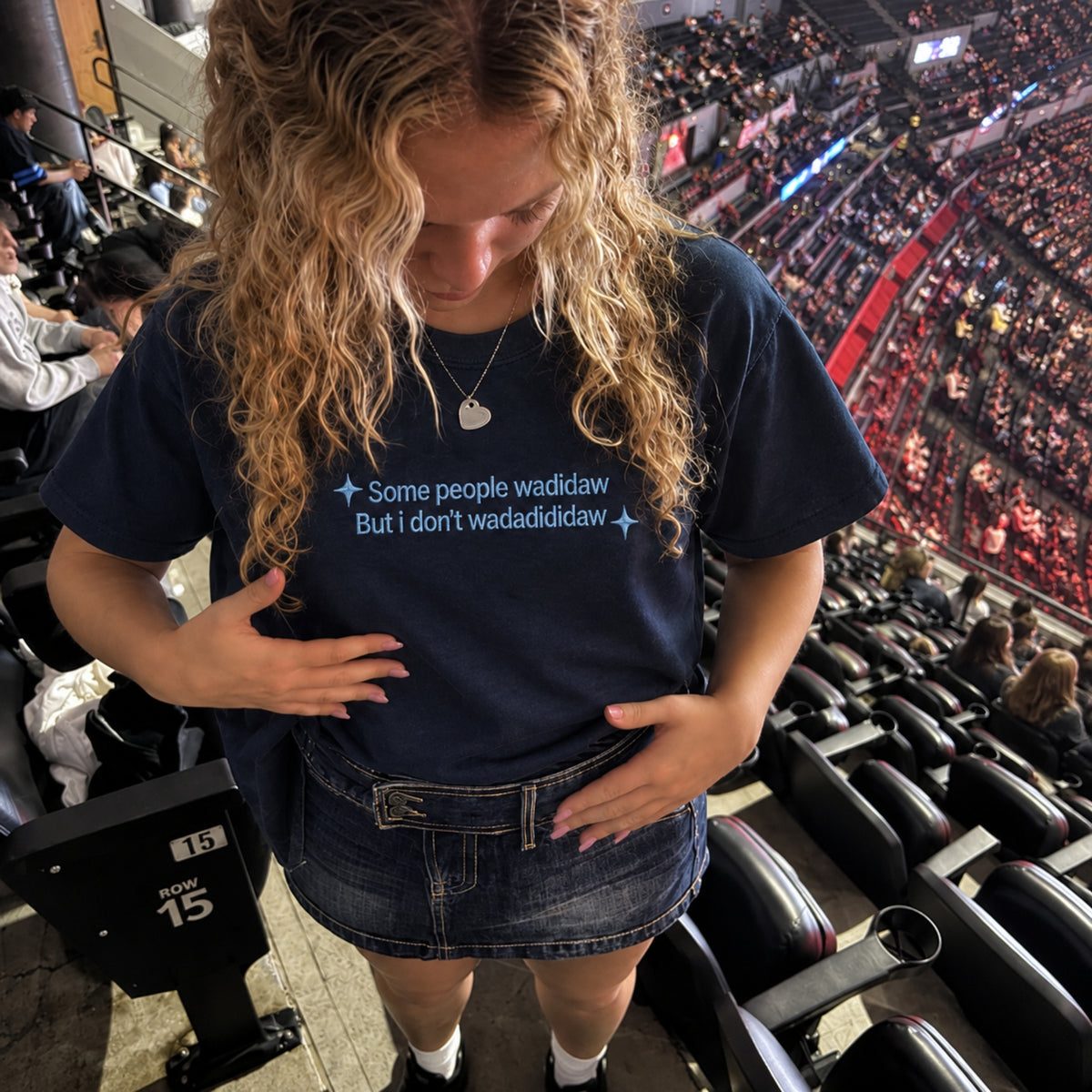 A woman in a crowded arena wearing a navy blue graphic t-shirt with the text 'Some people wadidaw But i don't wadidididaw' in light blue, paired with a denim mini skirt.