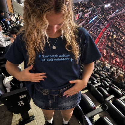 A woman in a crowded arena wearing a navy blue graphic t-shirt with the text 'Some people wadidaw But i don't wadidididaw' in light blue, paired with a denim mini skirt.
