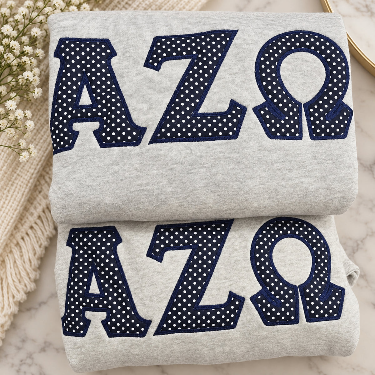 Two folded light heather gray sweatshirts stacked on a marble surface, each featuring large navy letters with white polka dots spelling AZΩ. A cream knit blanket and small white flowers are placed beside them as styling props.