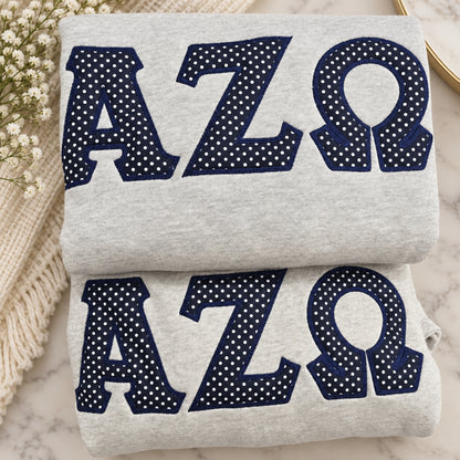 Two folded light heather gray sweatshirts stacked on a marble surface, each featuring large navy letters with white polka dots spelling AZΩ. A cream knit blanket and small white flowers are placed beside them as styling props.