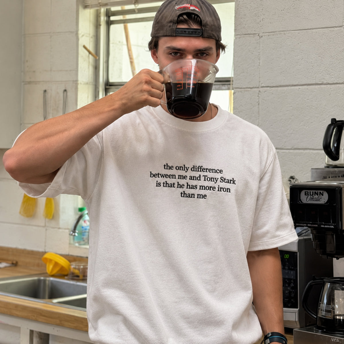 A man in a kitchen wearing a white graphic t-shirt with black text that reads 'the only difference between me and Tony Stark is that he has more iron than me'. He is holding a glass mug of coffee.